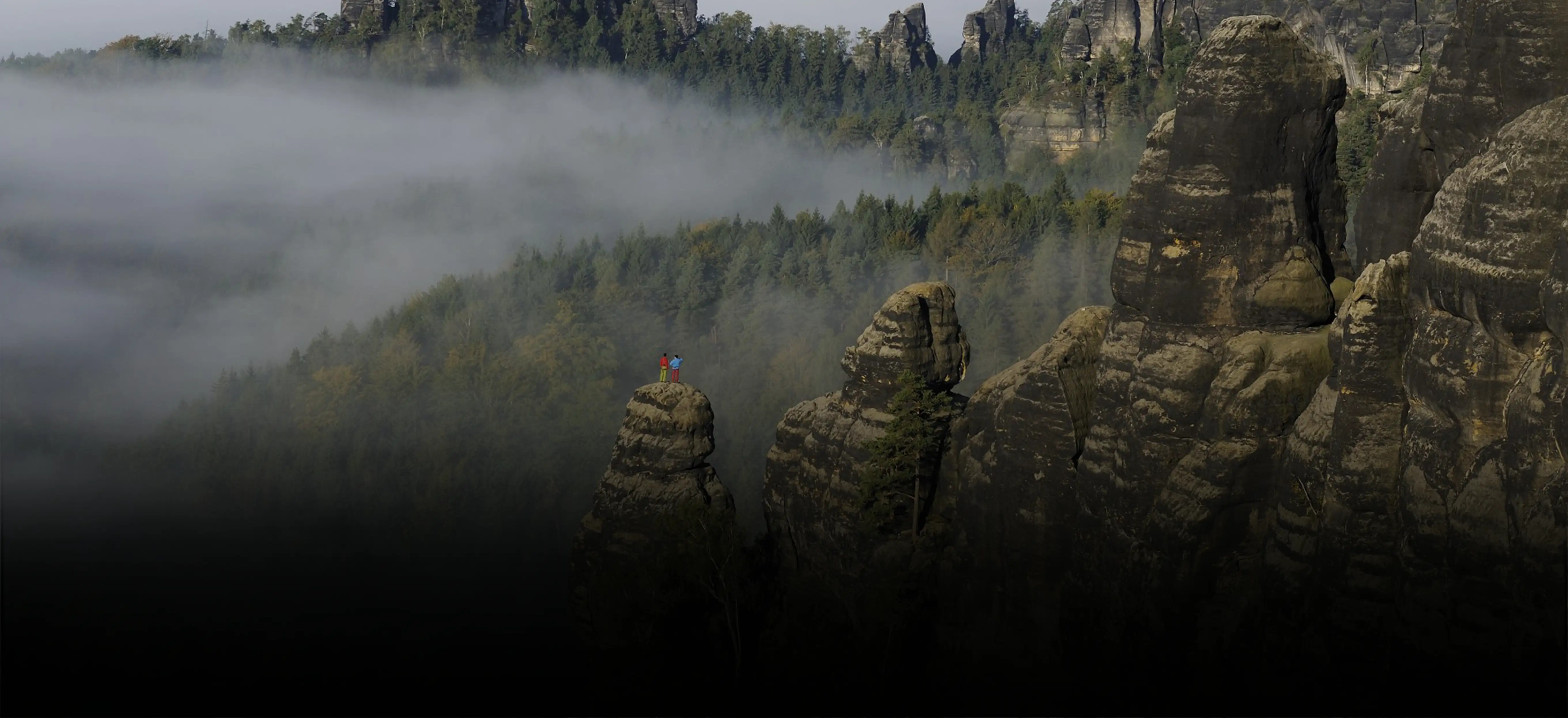 Zwei Wanderer blicken von einem Felsen auf die nebelverhangene Sächsische Schweiz